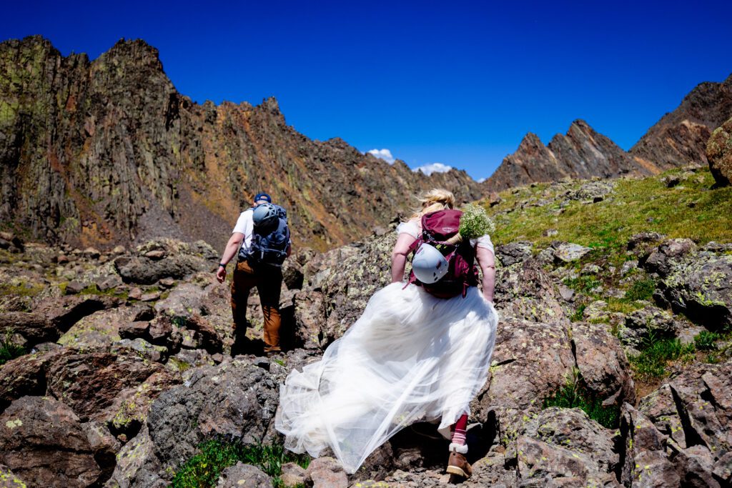A bride and a groom hiking Wetterhorn peak near Lake City Colorado with their helmets attached to their packs in preparation for a technical mountain hike