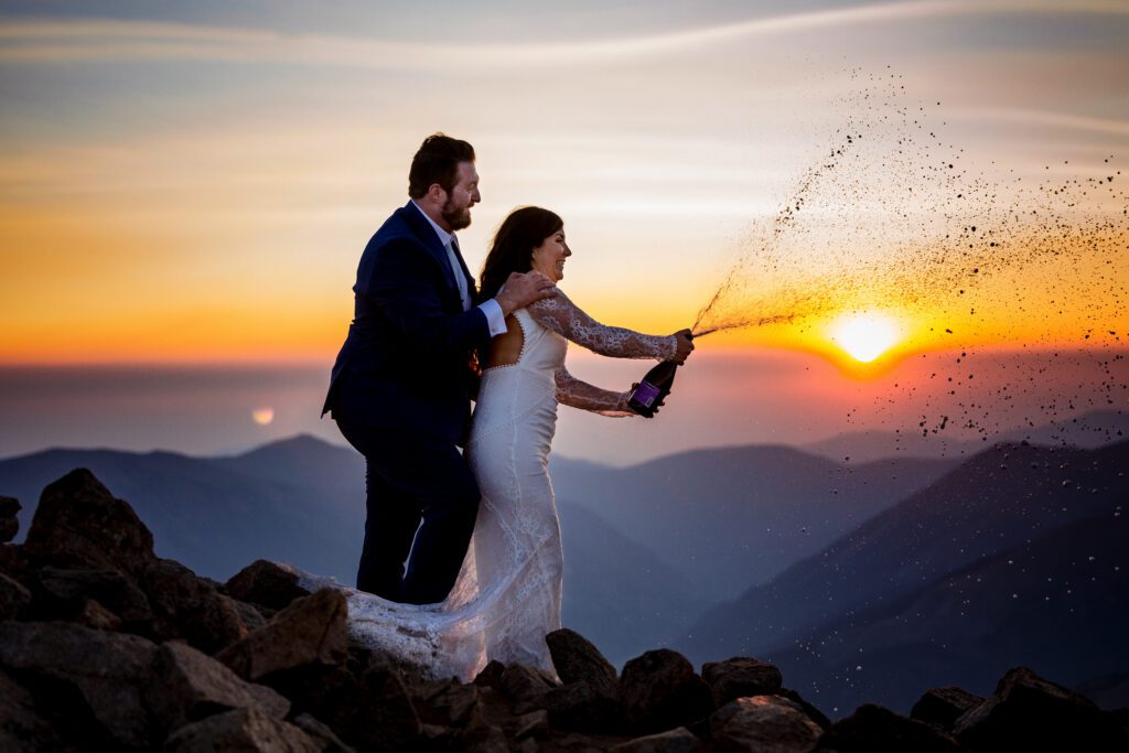 A bride and a groom at the top of Mt Sniktau in Colorado at sunrise popping a bottle of champagne in celebration