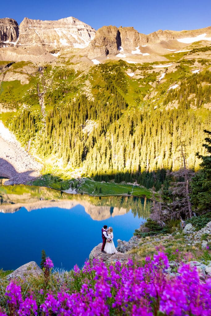 A couple embracing on a rock overlooking Lower Blue Lakes in Ridgway, Colorado with vibrant purple flowers in the foreground