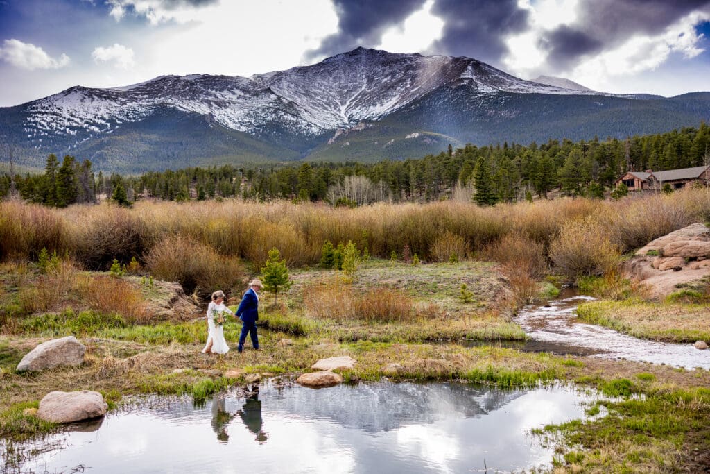 A bride and groom holding hands walking in front of an alpine lake in Estes Park with Mt. Meeker in the background on a spring day