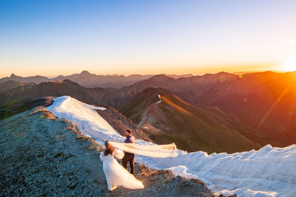 A bride and a groom on the top of Handies Peak at sunrise for their June elopement.  There is some snow still lingering at the summit and mountain peaks in the distance.